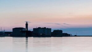 A waterfront view with a prominent lighthouse and several buildings, likely part of a historic or industrial area. - Alexander's Nest (Trieste)