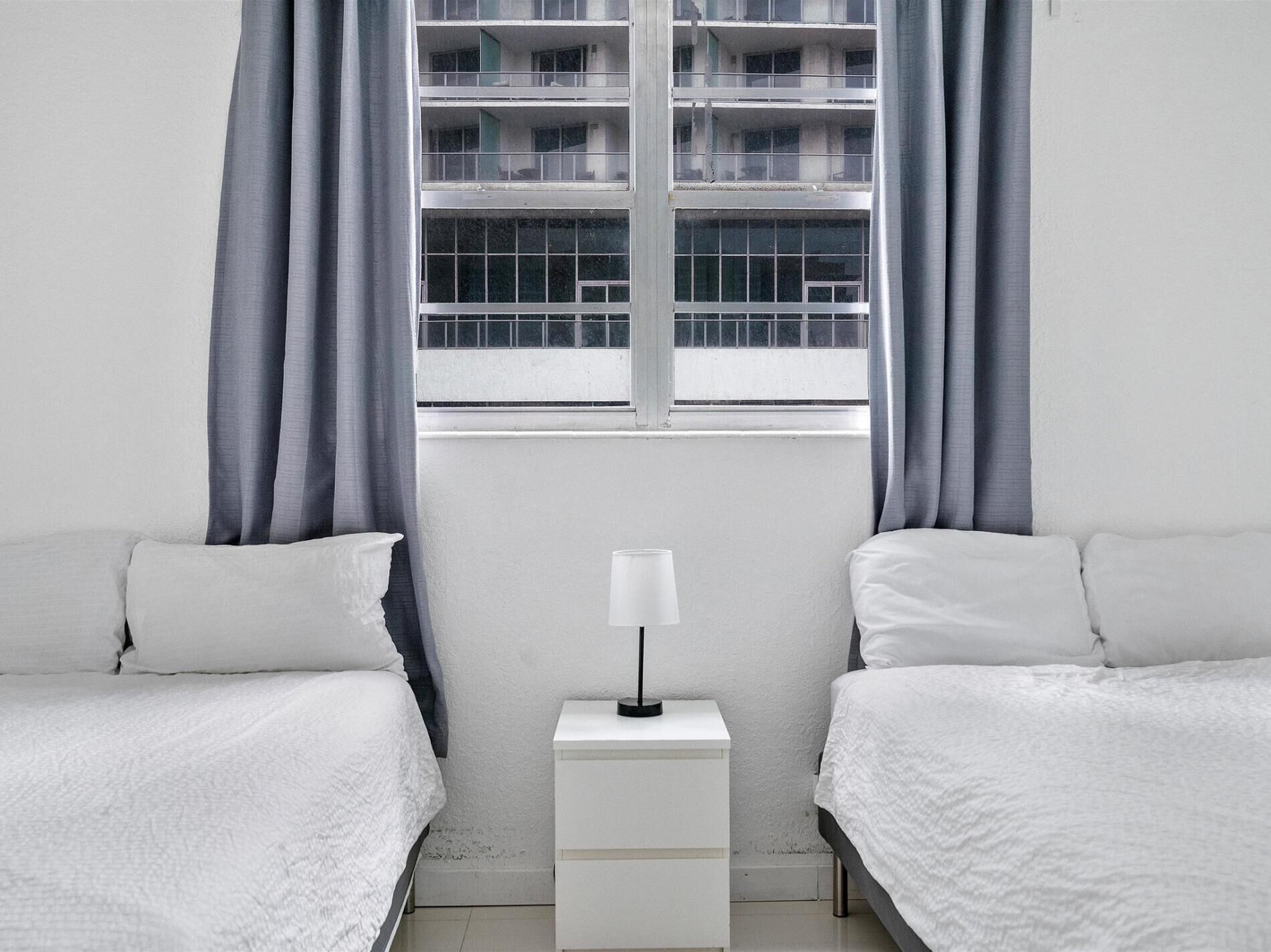 A minimalist bedroom with two beds, a nightstand, and a window with curtains.