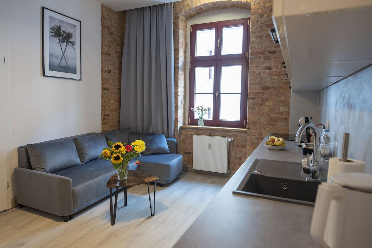 Ein modernes Wohnzimmer mit einem grauen Sofa, einem kleinen Tisch mit Blumenvase und einer Küchenzeile mit Spüle und Fenster mit Aussicht.