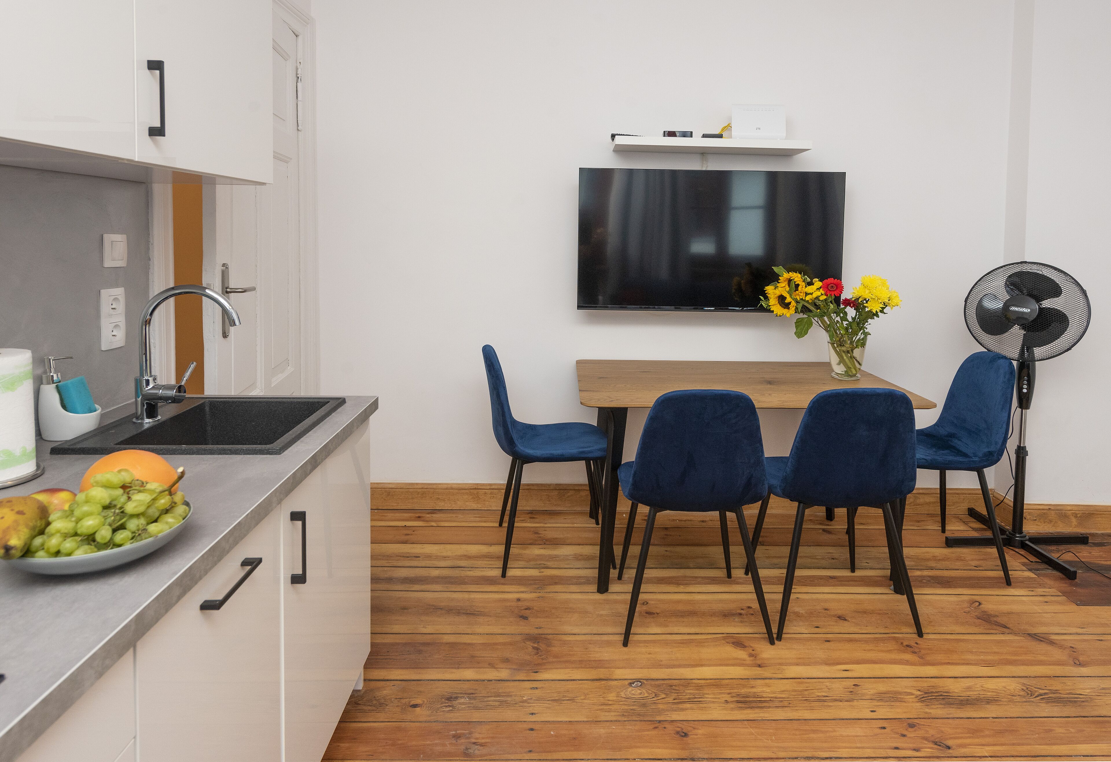 Eine moderne Küche mit einem Holztisch, blauen Stühlen, einem an der Wand befestigten Fernseher und einem Ventilator auf der rechten Seite.