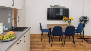A modern kitchen with a wooden dining table, blue chairs, a TV mounted on the wall, and a fan on the right.