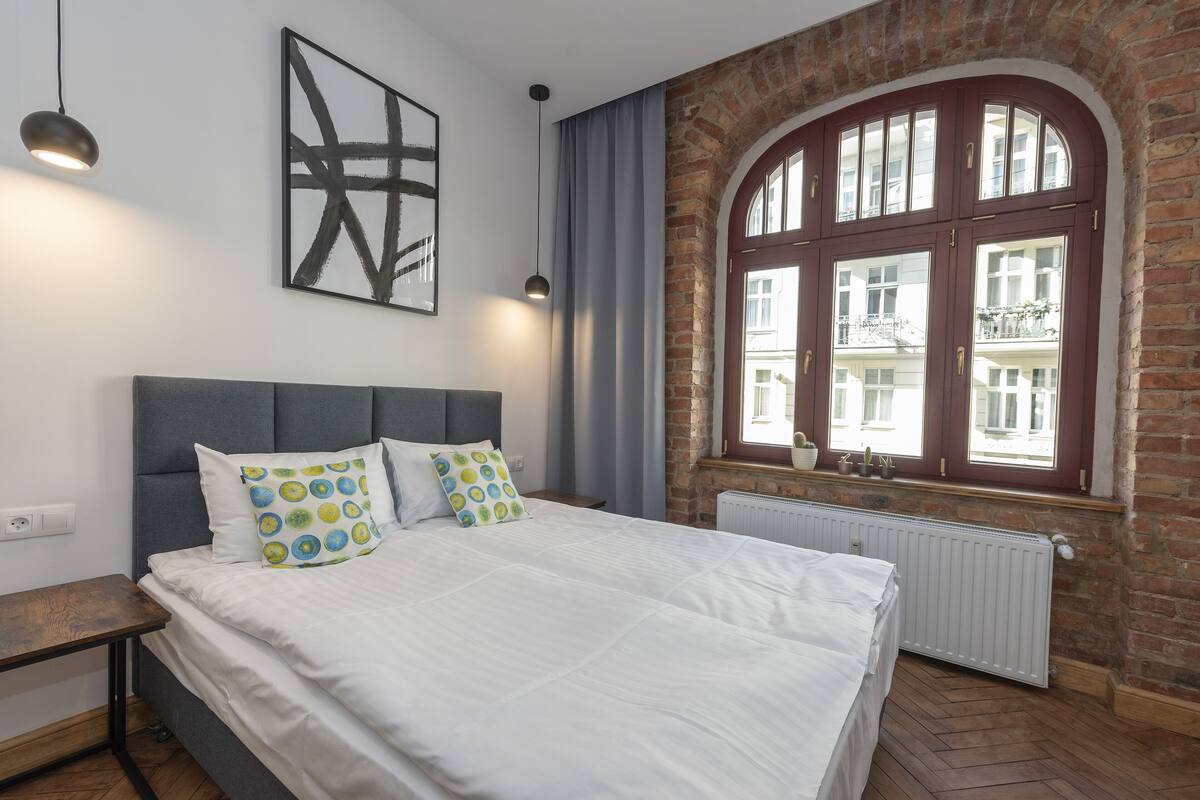 Une chambre à coucher avec un grand lit, une tête de lit, une fenêtre avec des rideaux, un radiateur et un mur en briques.