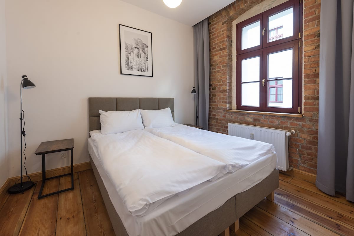 Ein Schlafzimmer mit einem großen Bett, einem Nachttisch, einer Stehlampe, einer Heizung und einem Fenster mit Blick auf ein Gebäude.