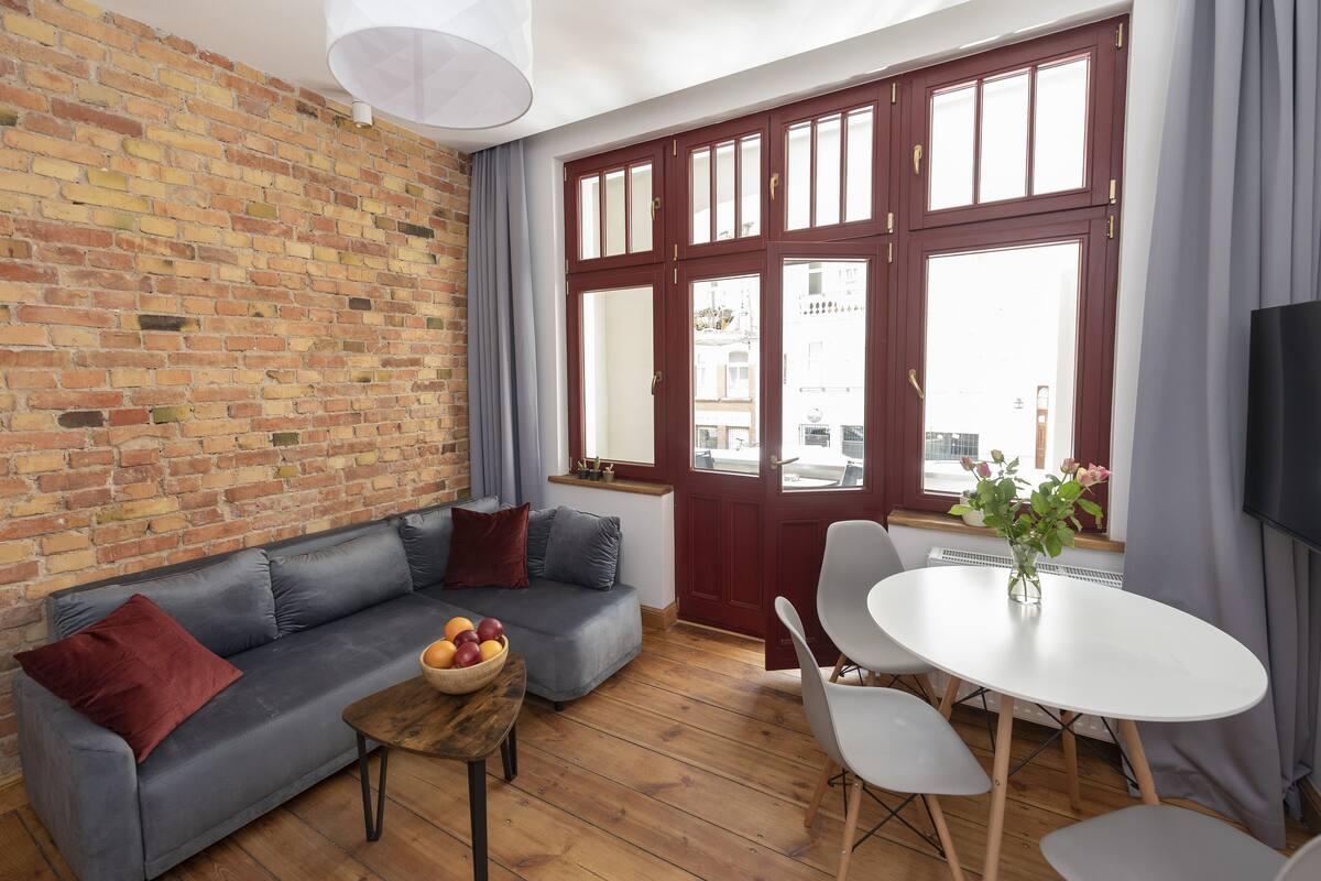 Un salon avec un canapé gris, une table basse en bois sur laquelle se trouvent des fruits, une table blanche avec des chaises et un mur en briques.