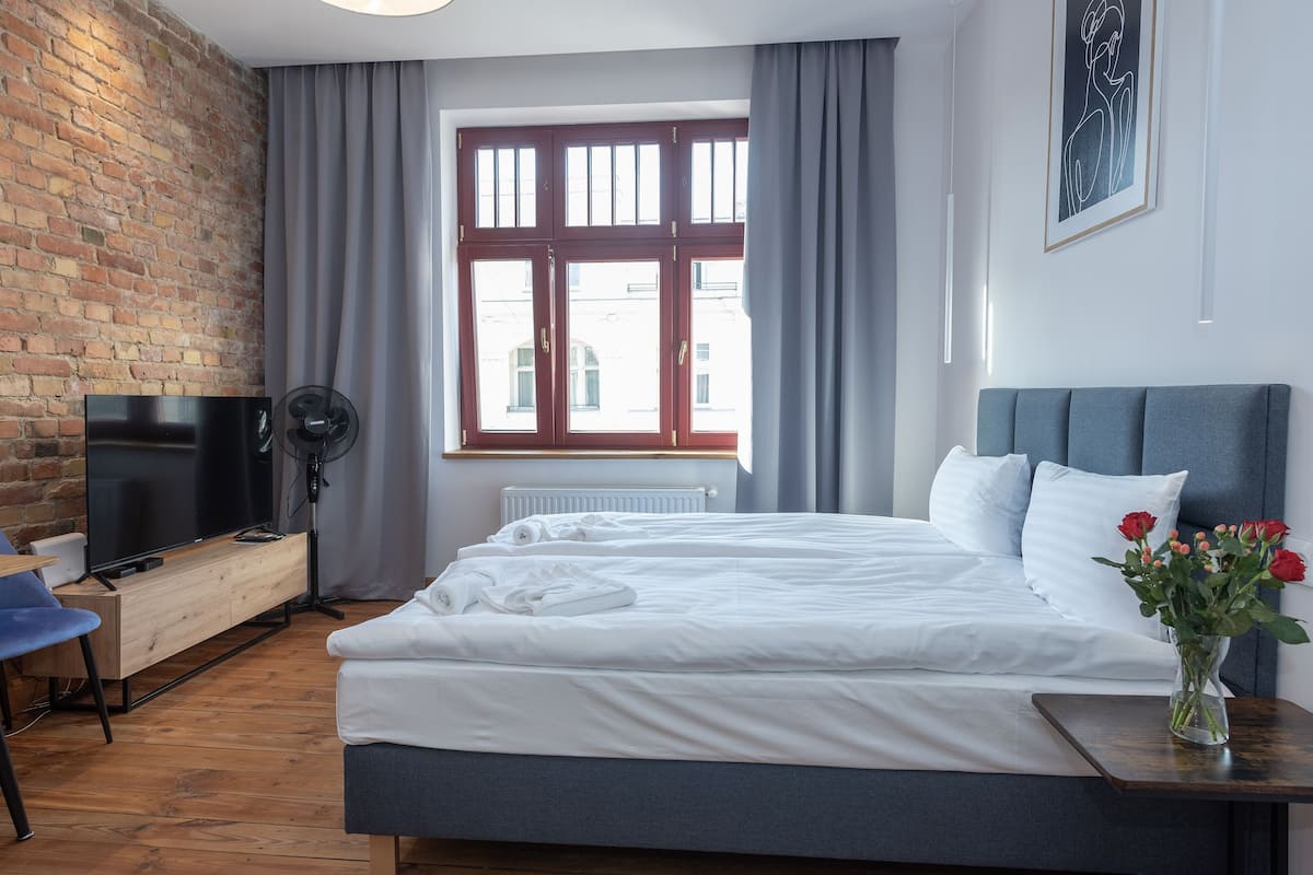 Une chambre d’hôtel moderne avec un mur en briques, une grande fenêtre avec des rideaux, un téléviseur à écran plat, un bureau en bois, un lit avec du linge de maison blanc et un vase avec des fleurs.