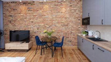 A compact kitchen with a brick wall, wooden flooring, a dining table with chairs, a TV, and modern cabinetry.