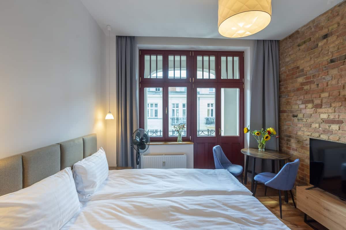Ein Hotelzimmer mit einem großen Bett, einem Fernseher, einem kleinen Tisch mit Stühlen und einem Fenster mit Blick auf Gebäude.