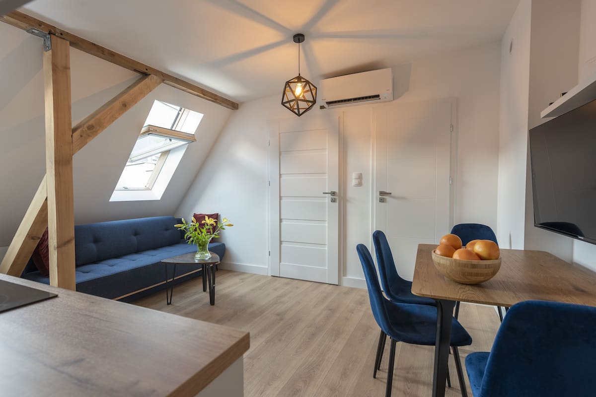 Ein modernes Loft-Apartment mit schrägverdeckter Decke, einem Essbereich mit Holztisch und blauen Stühlen, einem blauen Sofa und einem kleinen Beistelltisch mit Blumenvase.
