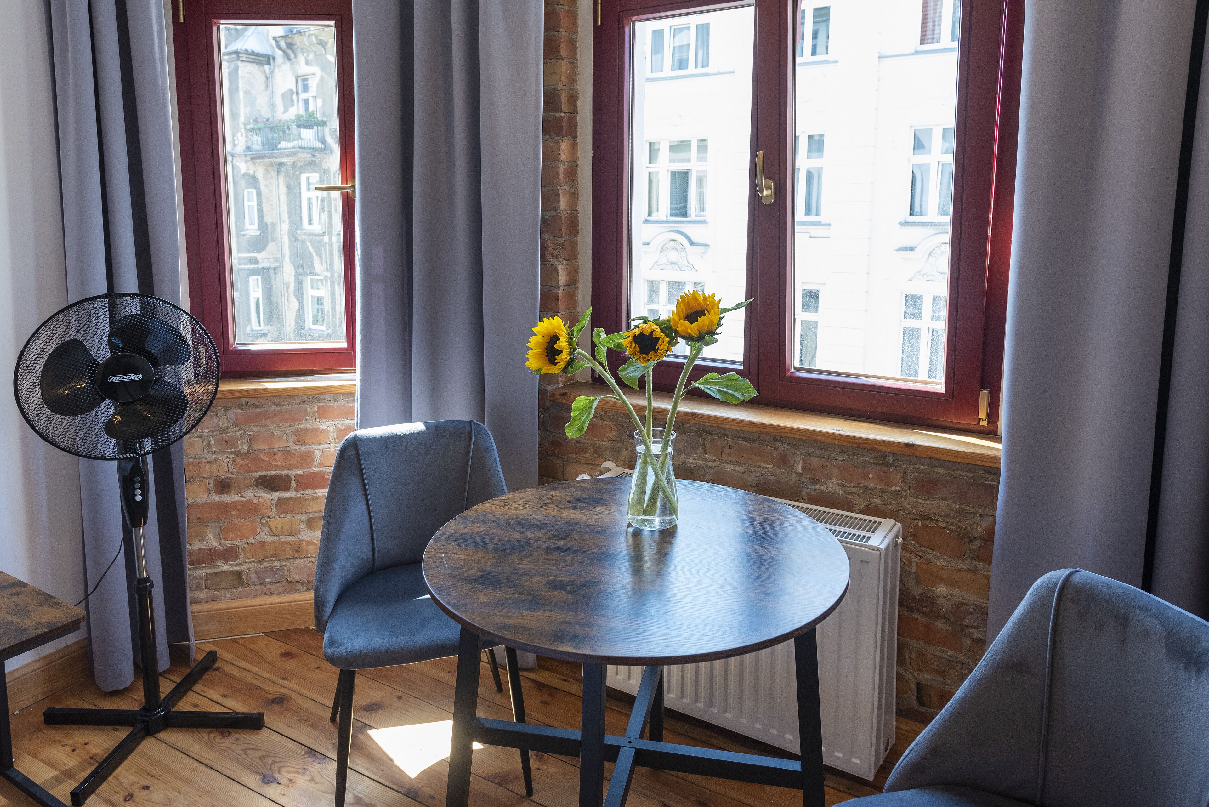 Una habitación con una mesa redonda, dos sillas, un jarrón con girasoles, un ventilador de pie y vistas a edificios a través de una ventana con marcos rojos.
