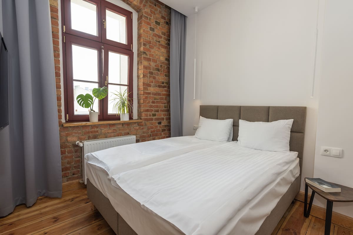 Ein Schlafzimmer mit Doppelbett, Backsteinwand, einem Fenster mit Vorhängen und einer Topfpflanze.