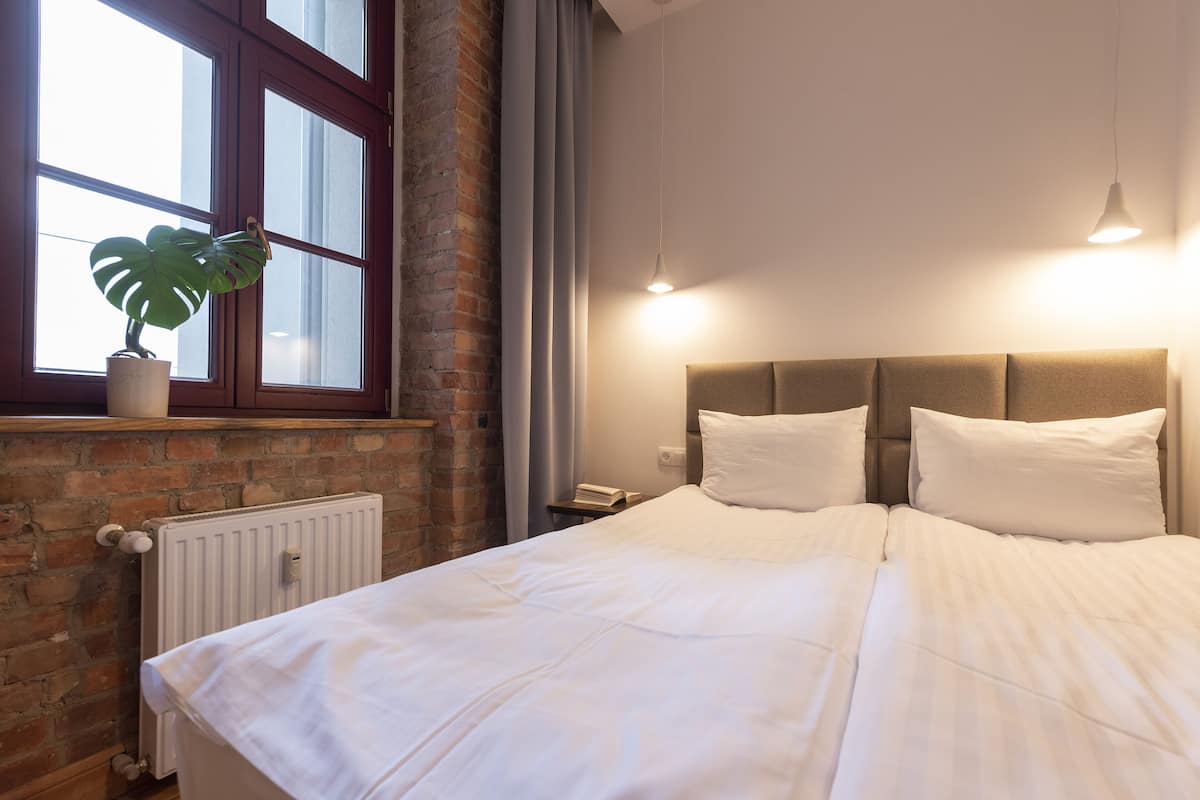 Une chambre avec un grand lit, une plante en pot, une fenêtre avec des rideaux et un mur en briques.