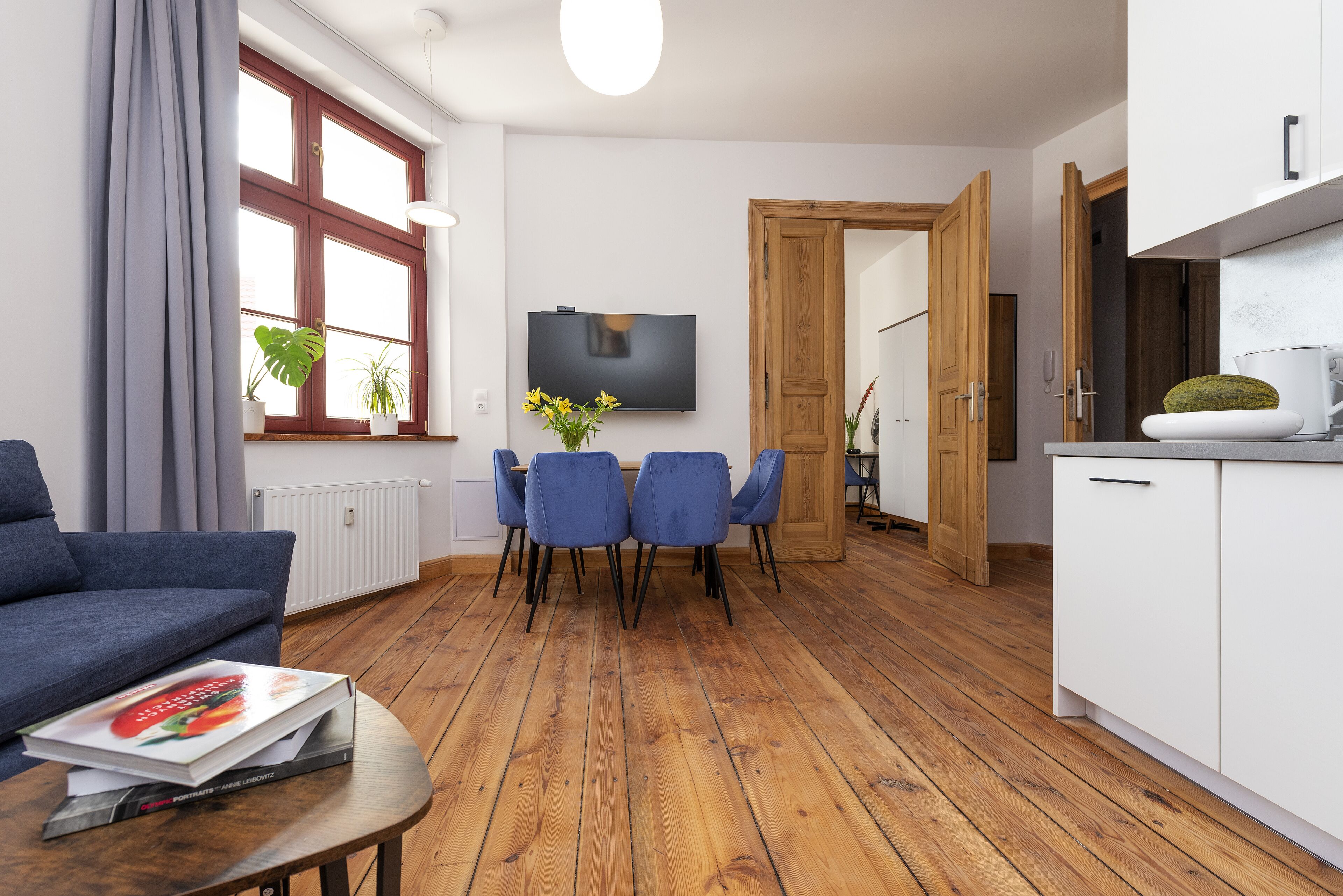A modern living room with a blue sofa, a dining table with four chairs, a TV mounted on the wall, and wooden flooring.
