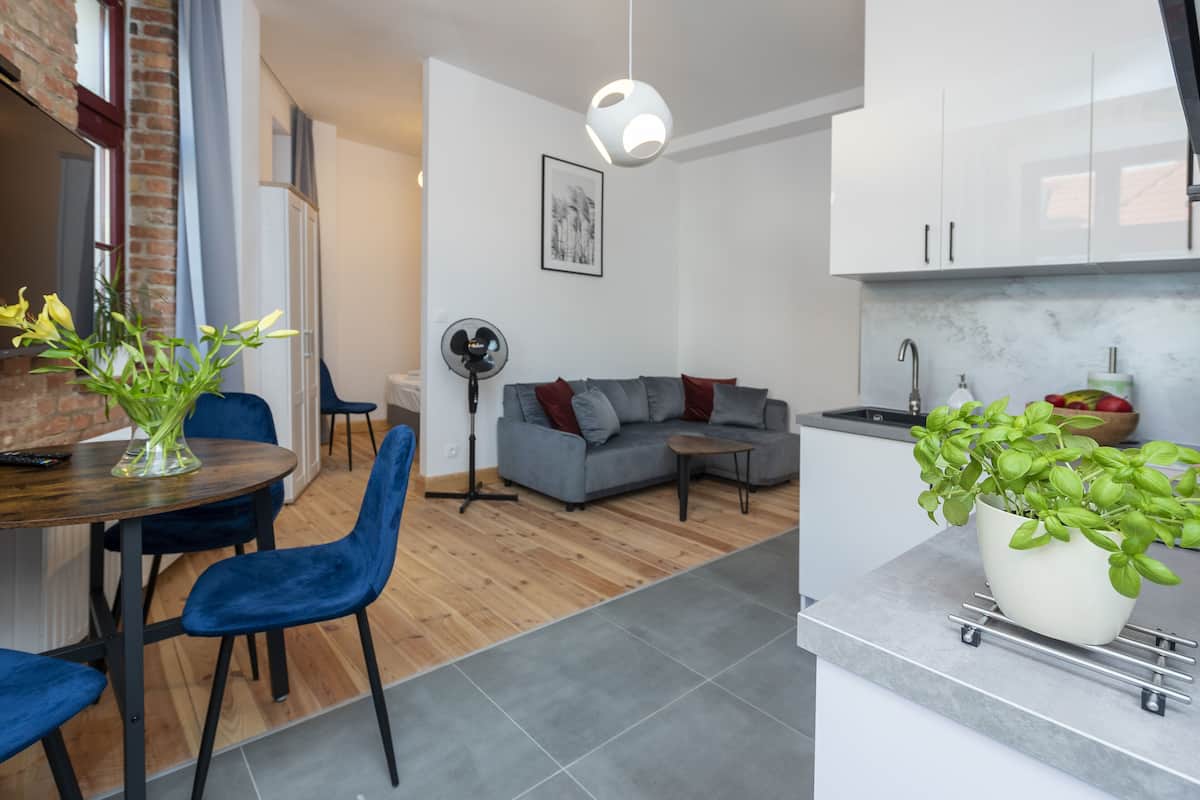 Ein modernes Wohnzimmer mit grauem Sofa, blauen Sesseln, einem Esstisch mit Blumenvase sowie einer Küchenzeile mit weißen Schränken und einem Topf mit Basilikum.
