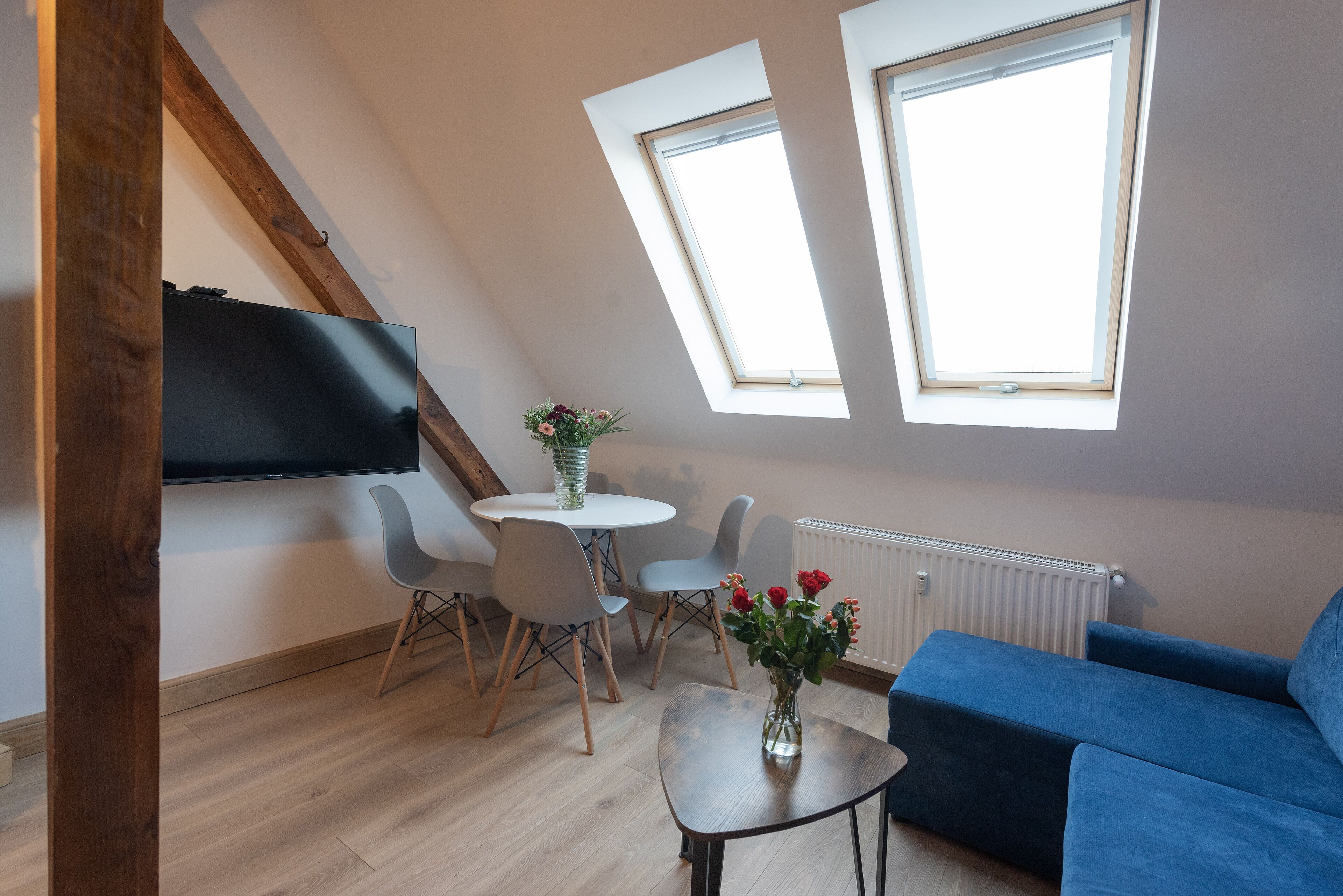 Un salón moderno con un sofá azul, una mesita redonda con un florero, y un televisor montado en la pared. Hay dos tragaluces que proporcionan luz natural y una zona de comedor con sillas y una mesa.