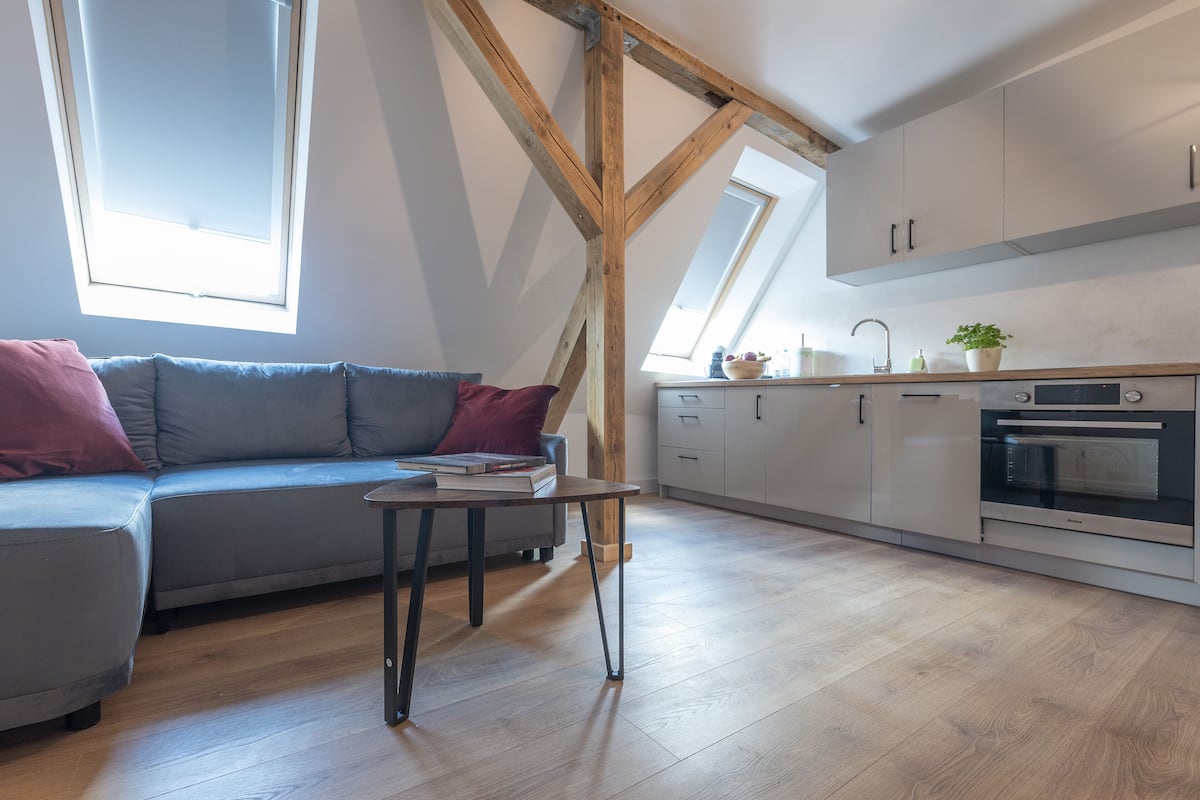 Eine moderne Loft-Wohnung mit Schlafgalerie, einem grauen Sofa mit roten Kissen, einem kleinen Tisch und einer Küche mit weißen Schränken und einem eingebauten Backofen.