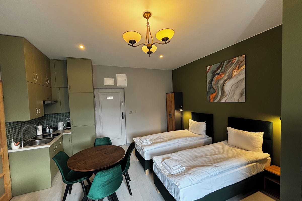 Ein kompaktes Hotelzimmer mit zwei Einzelbetten, einer kleinen Küchenzeile, einem Essbereich mit Tisch und Stühlen sowie einer Deckenleuchte.