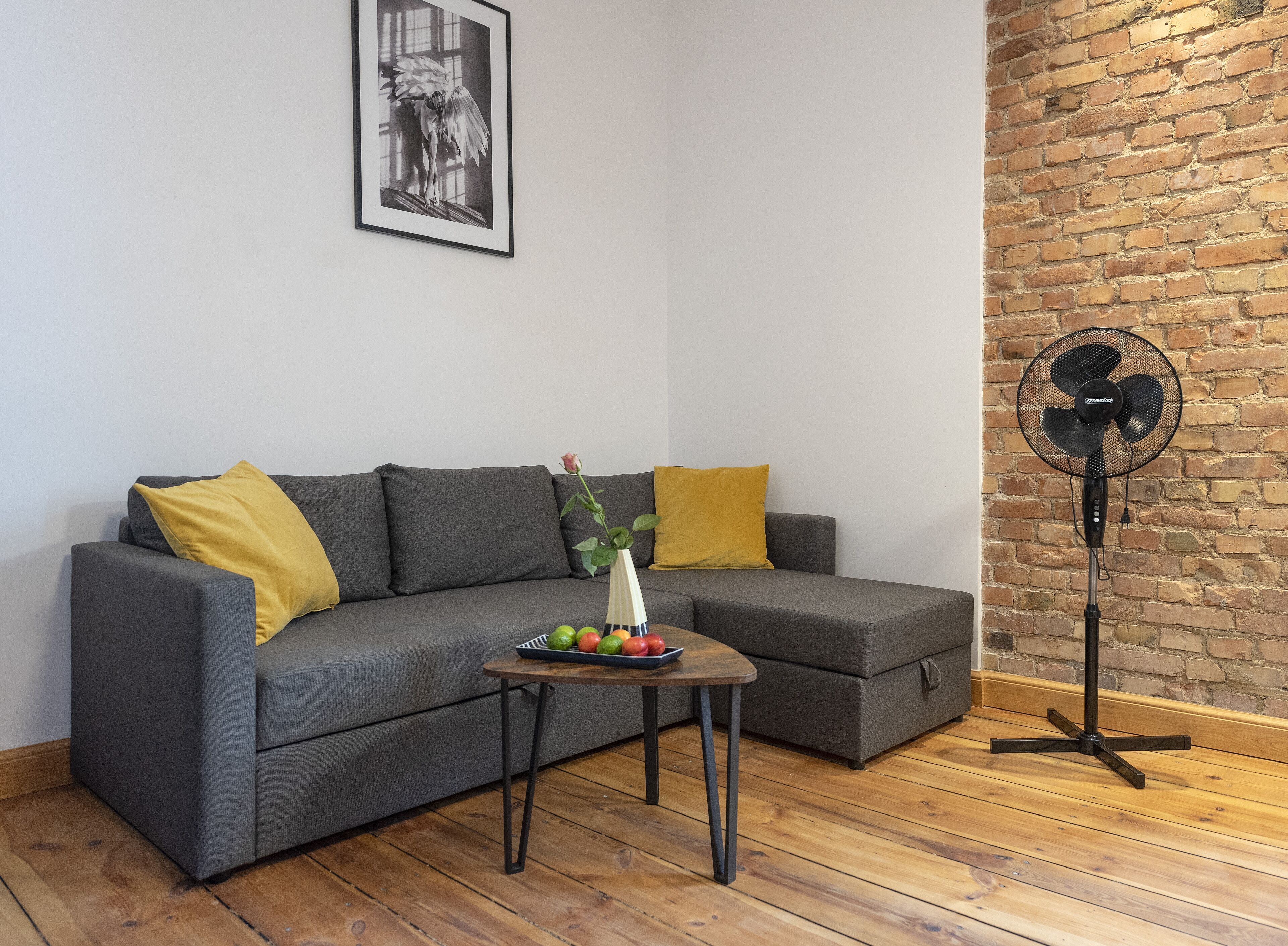 Uma sala de estar moderna com um sofá cinzento, almofadas amarelas, uma pequena mesa de centro de madeira com frutas e um ventilador de chão.