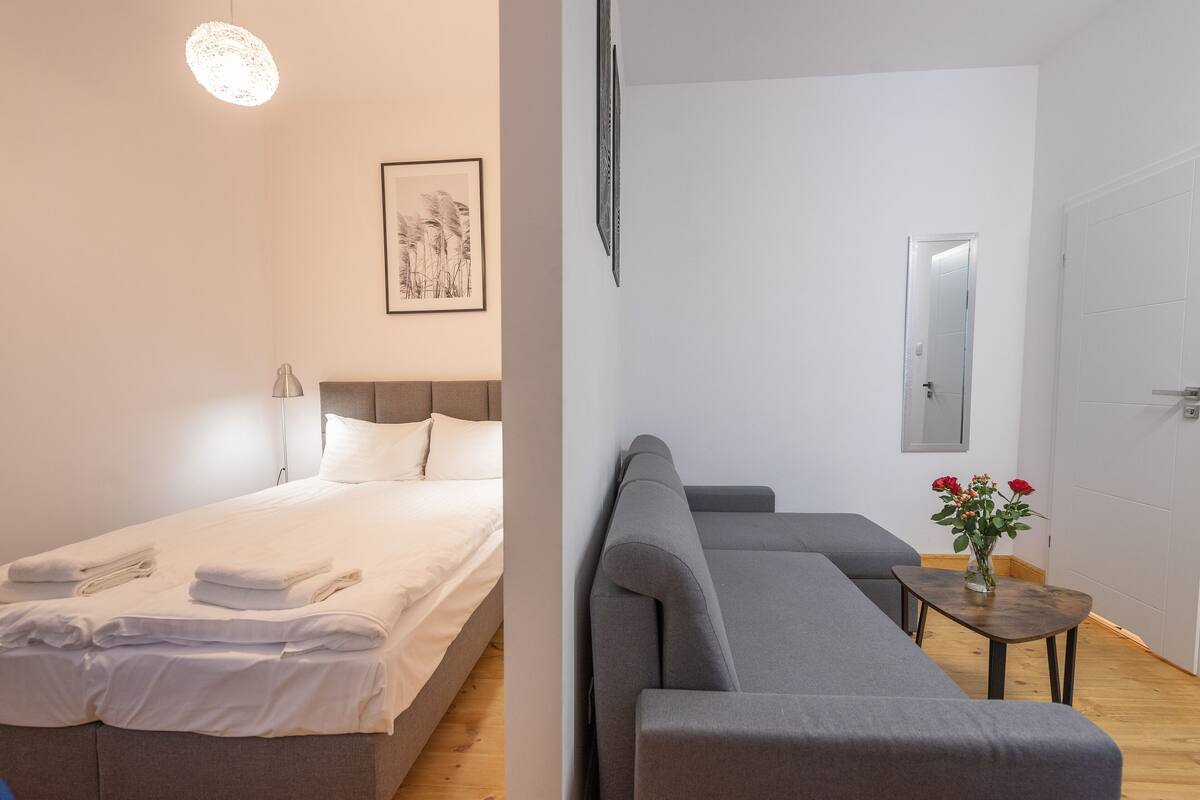 Ein Zimmer mit einem Bett, einem grauen Sofa, einem kleinen Tisch mit Blumen und einem gerahmten Bild an der Wand.