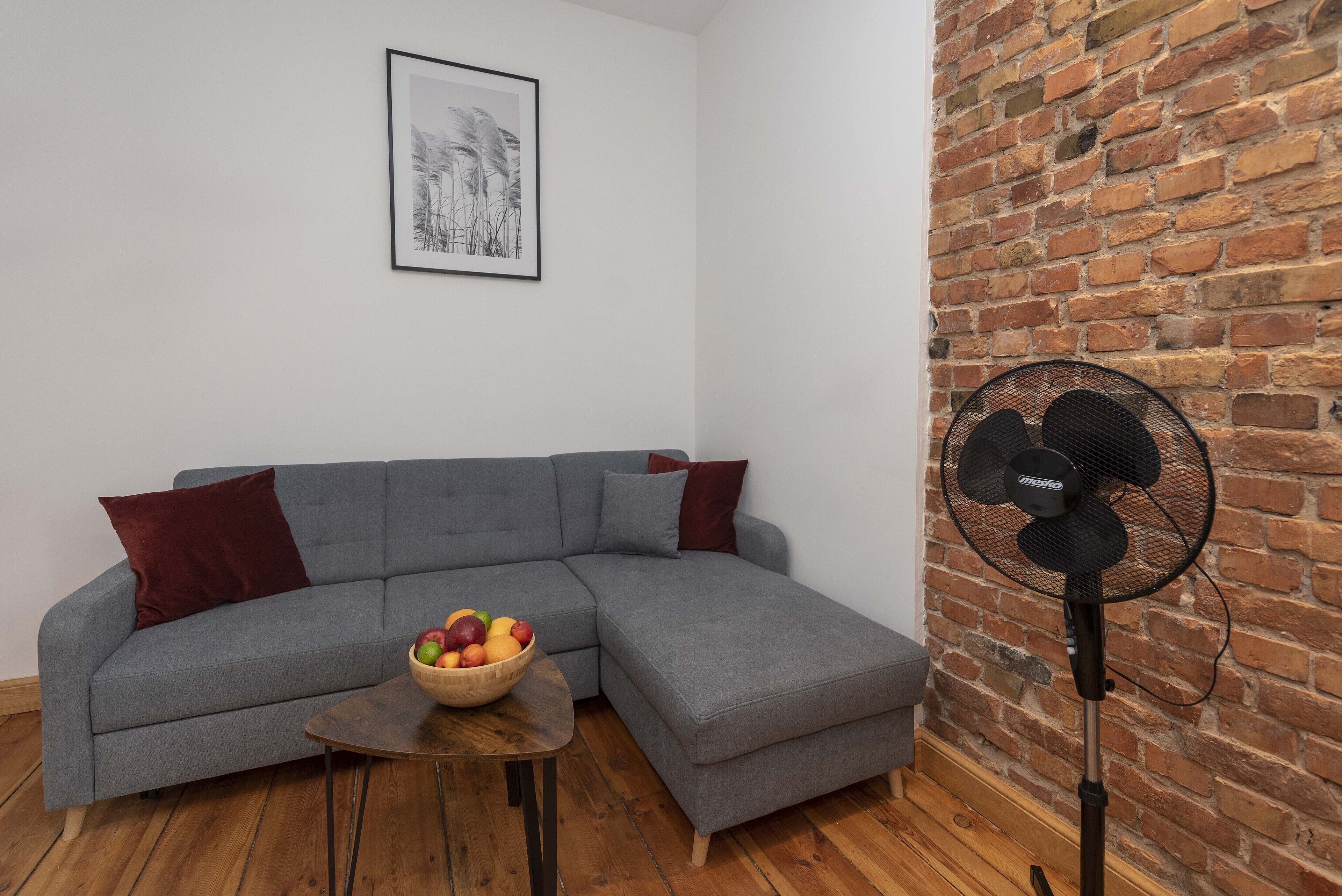 A living room with a grey sectional sofa, a wooden coffee table with a fruit bowl, a standing fan, and a brick wall.