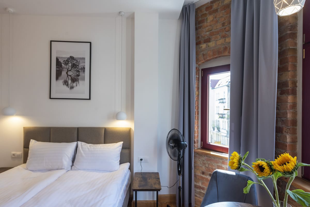 Une chambre d’hôtel avec un lit, un ventilateur sur pied, une chaise, une fenêtre avec des rideaux et un tableau encadré au mur.