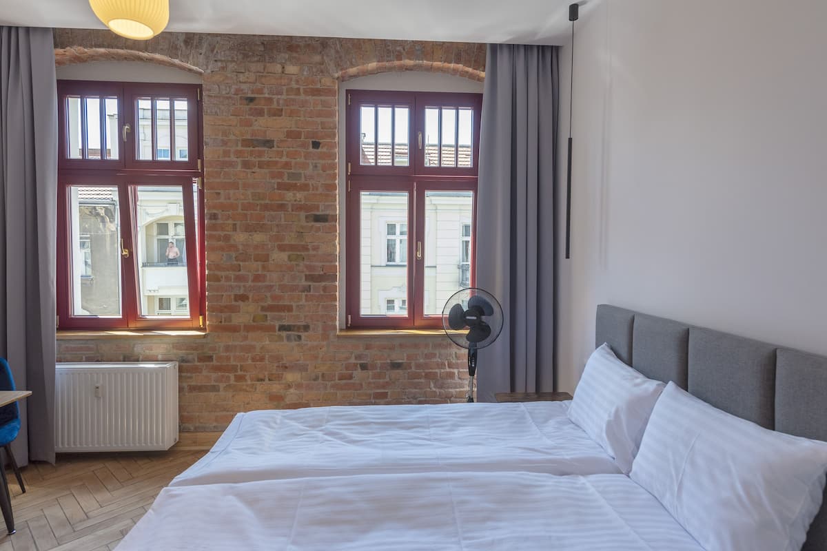 Une chambre avec un mur en briques, deux fenêtres avec des rideaux, un lit avec du linge de lit blanc, un ventilateur sur pied et un radiateur.