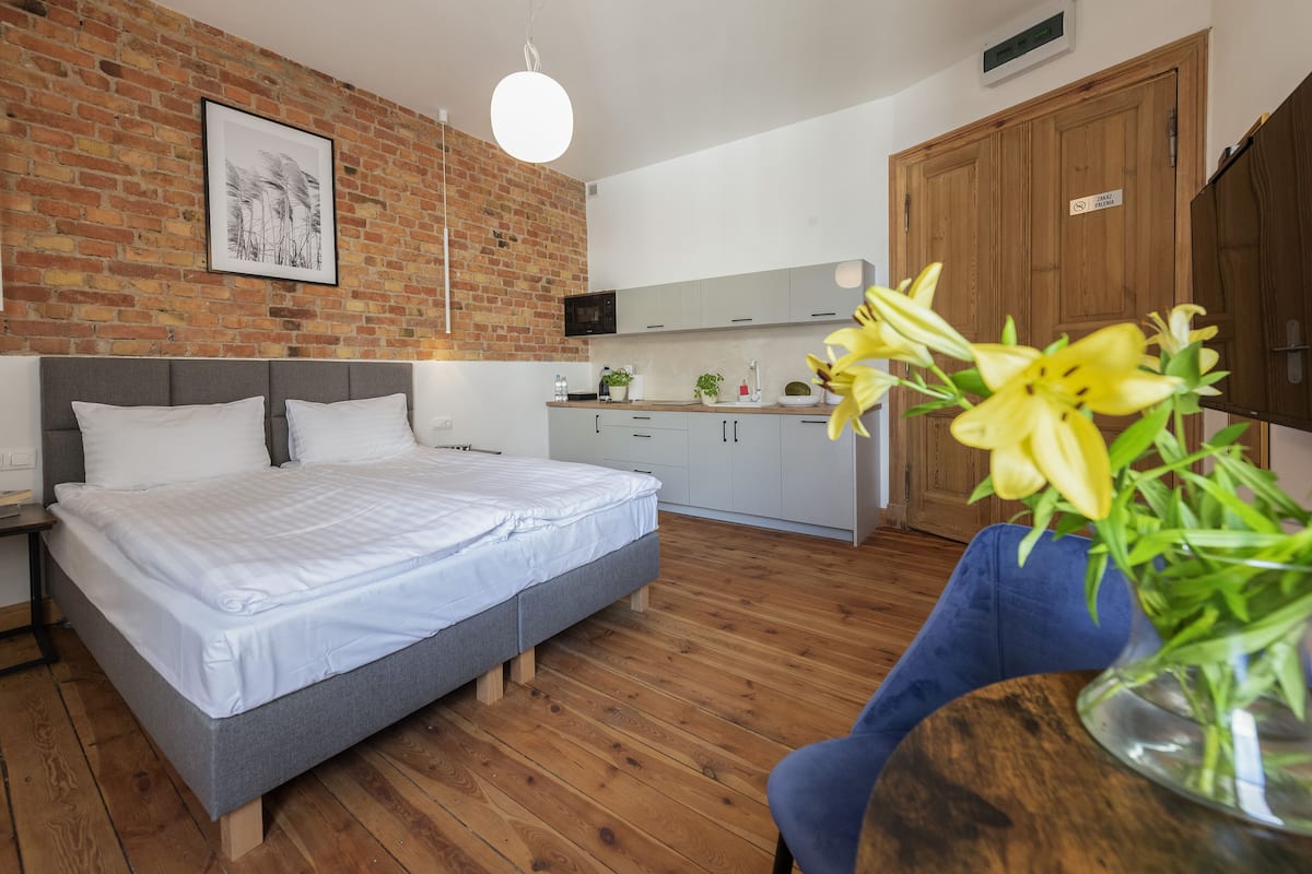 Une chambre à coucher avec un lit, un plancher en bois, un mur en briques, un coin cuisine avec des placards et une porte en bois.
