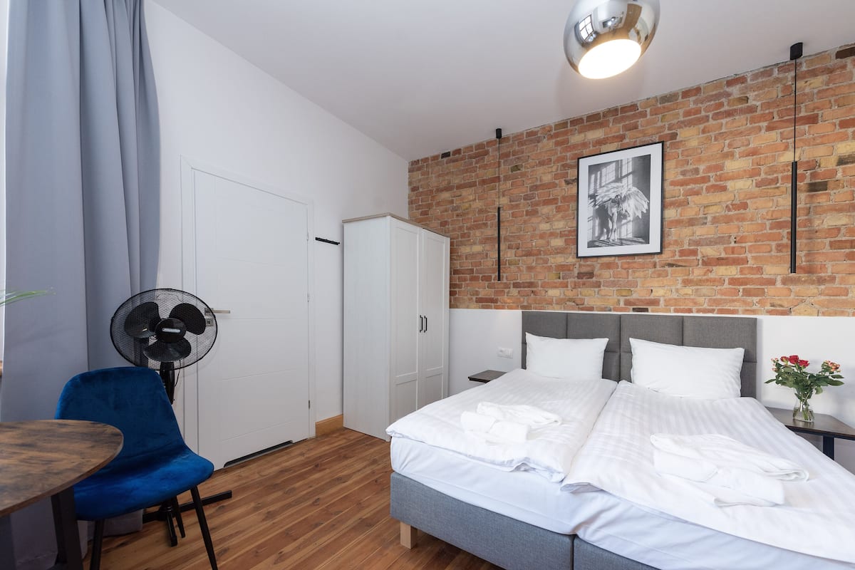 Une chambre d’hôtel avec un mur en briques, un lit avec du linge de maison blanc, une chaise bleue, un ventilateur et une petite table.