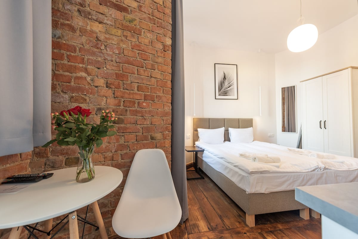 Une chambre d’hôtel moderne avec un mur en briques, un lit avec du linge de maison blanc, une table de chevet avec une télécommande et un vase rempli de fleurs.