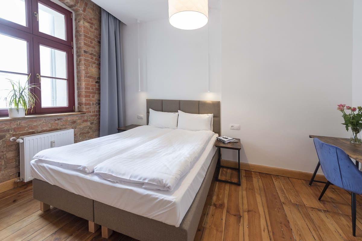 Un lit bien fait dans une pièce dont un mur est en briques, avec une fenêtre ornée de rideaux, un plancher en bois et un bureau avec une chaise.