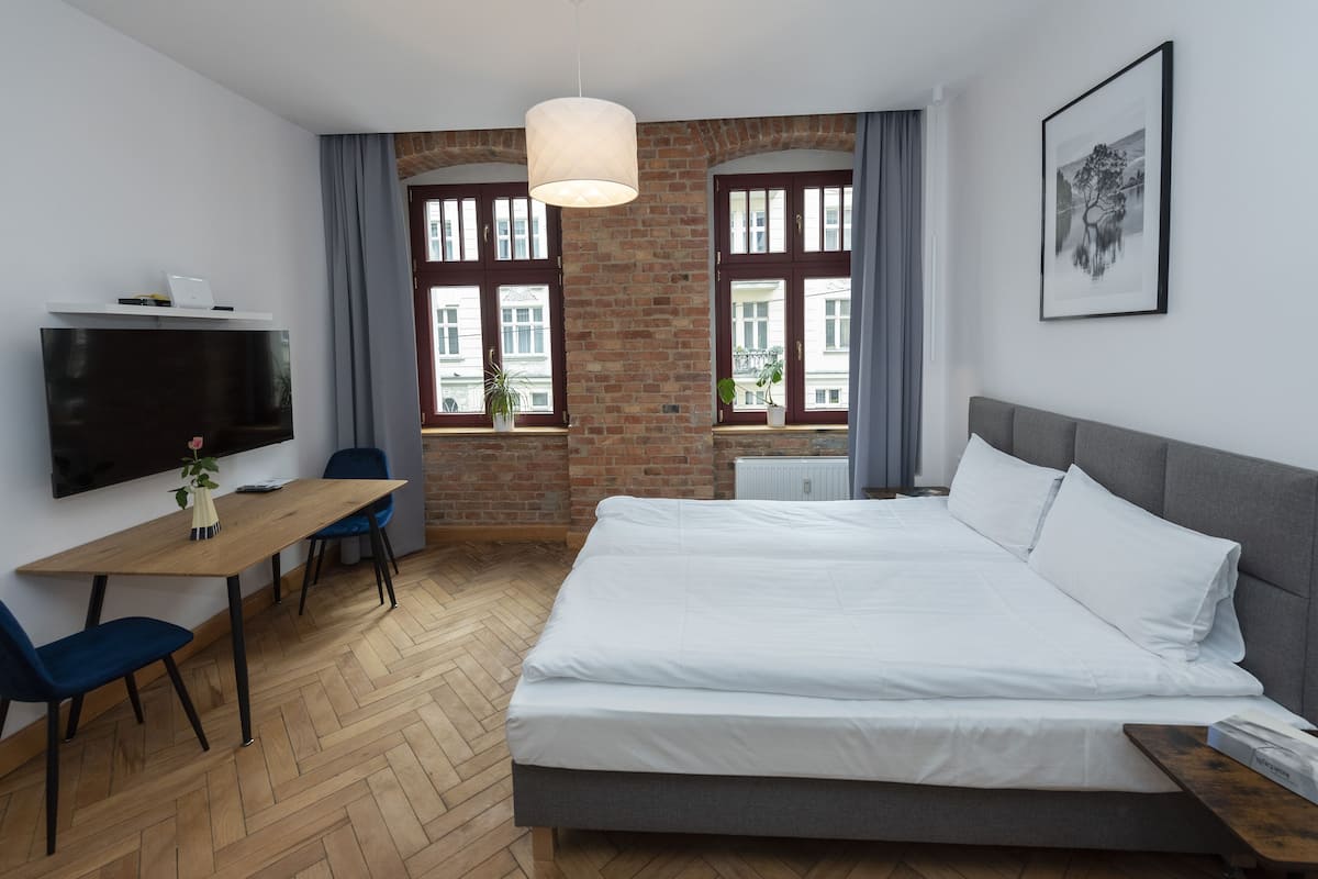 Une chambre d’hôtel moderne dotée d’un grand lit, d’un bureau avec un ordinateur portable, de deux chaises, d’une télévision et de grandes fenêtres avec des rideaux.