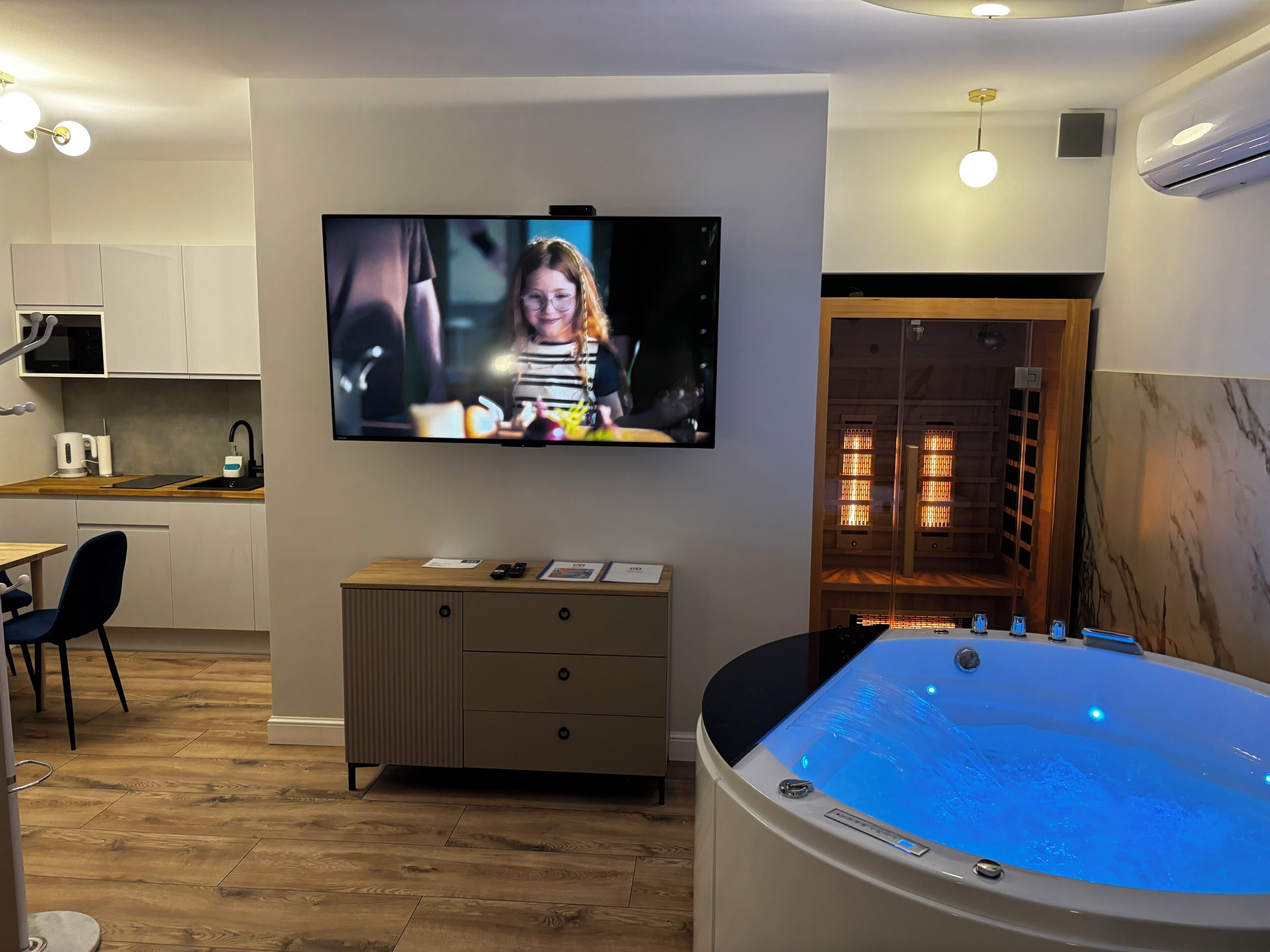Una habitación de hotel moderna con una televisión de pantalla plana montada en la pared, un mueble de madera con calefacción integrada, una bañera de hidromasaje con iluminación azul y una zona de cocina con microondas y hervidor.