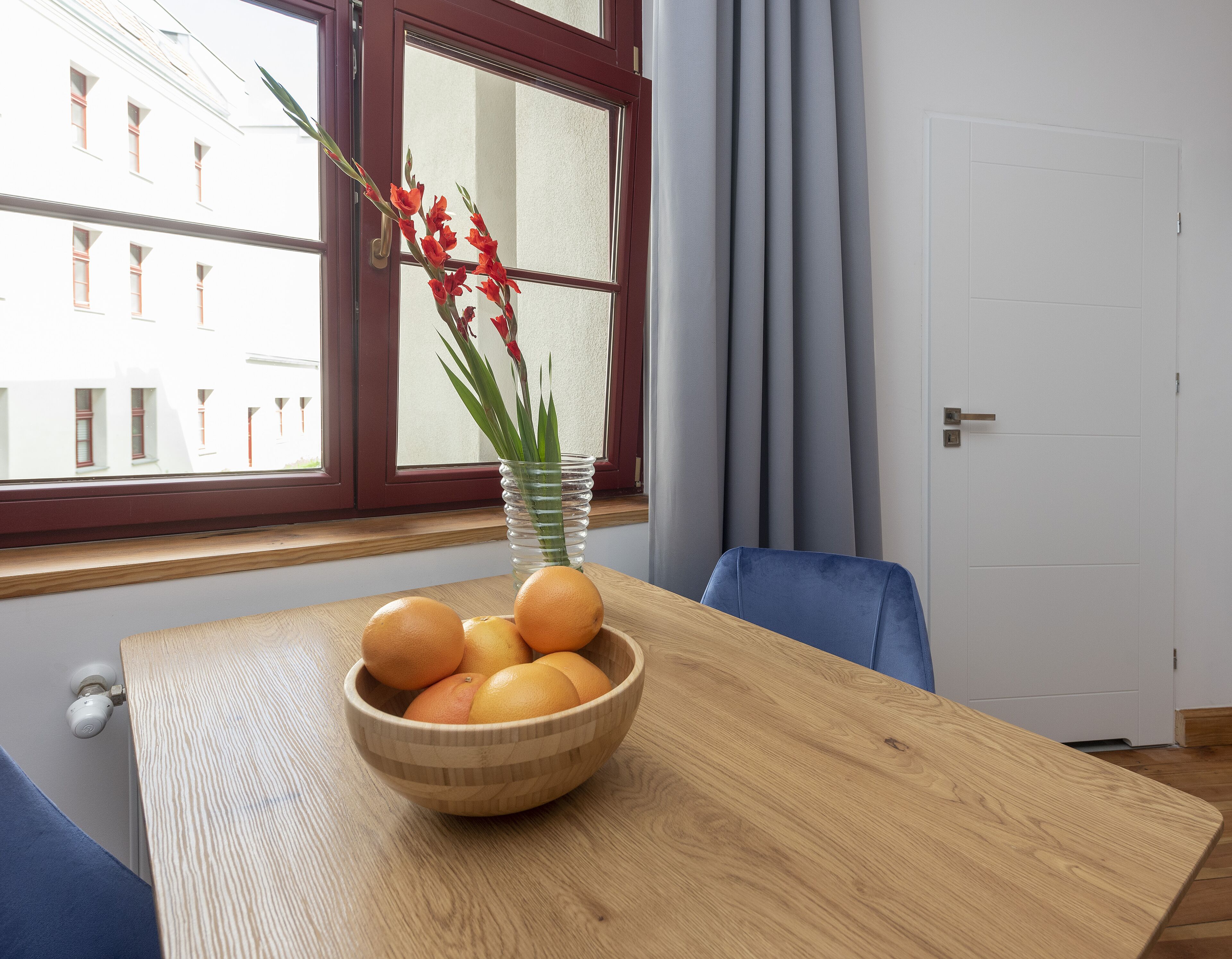 A wooden dining table with a bowl of oranges and a vase with flowers.
