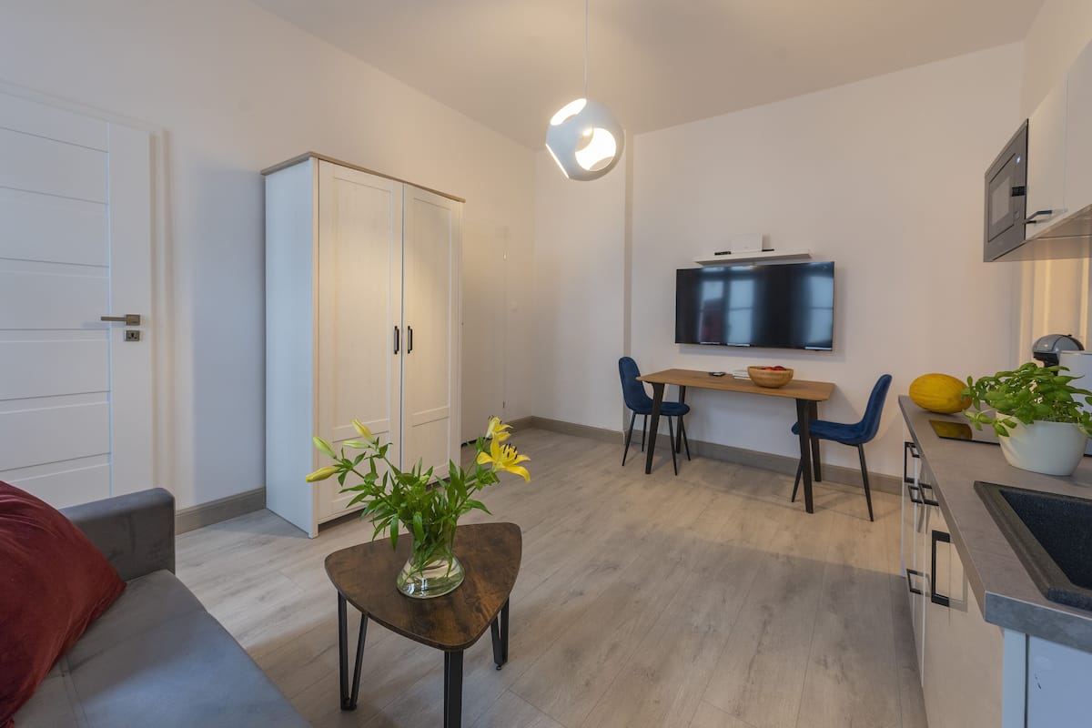Ein modernes Wohnzimmer mit einer Couch, einem Esstisch mit Stühlen, einem an der Wand befestigten Fernseher und einer Küchenzeile mit Spüle und Schränken.