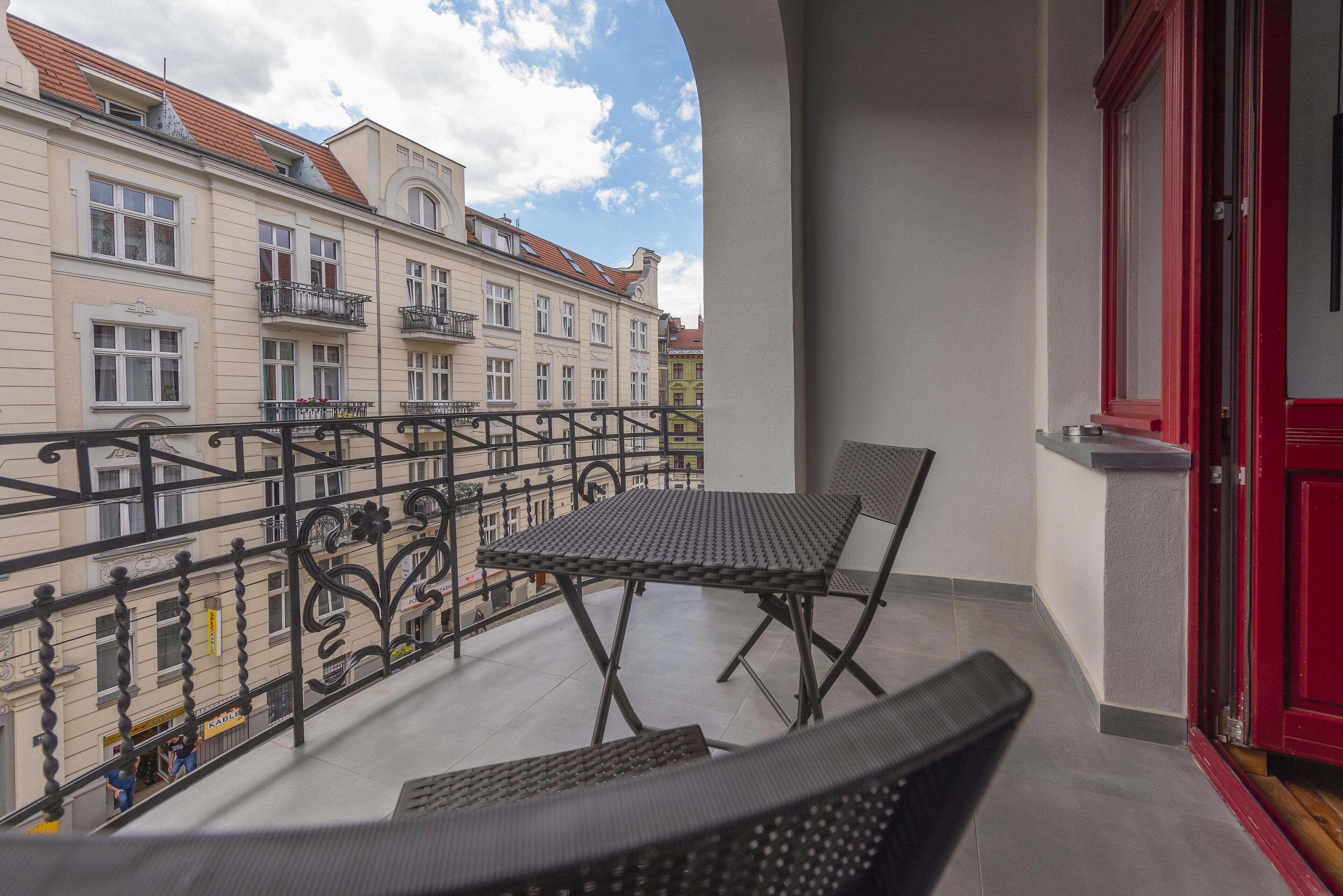 A balcony with a table and chairs, a red door, and a view of buildings with balconies.