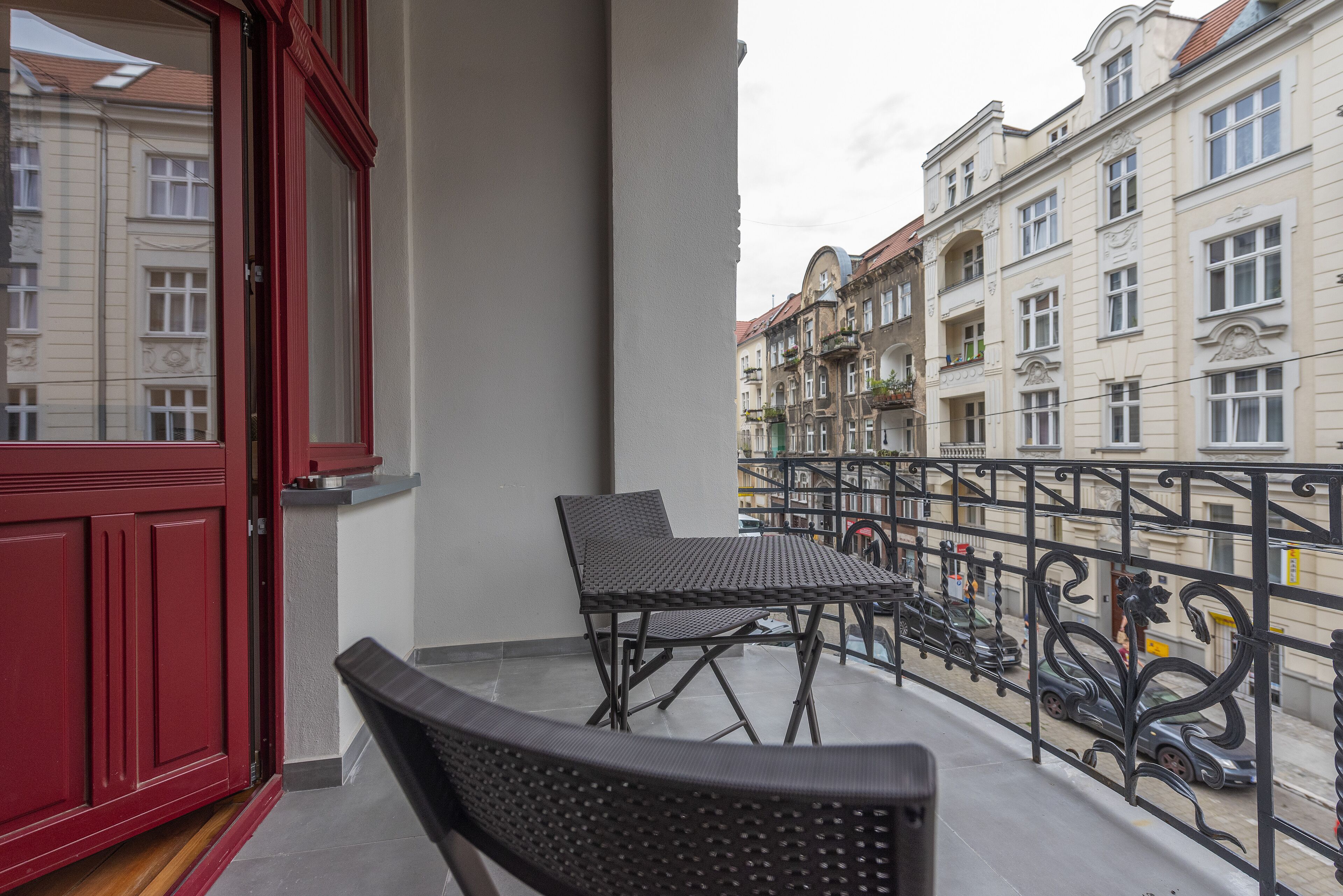 Un balcón con una puerta roja, una mesa y silla de metal negro, y vistas a edificios vecinos.