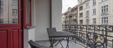 Un balcón con una puerta roja, una mesa y silla de metal negro, y vistas a edificios vecinos.