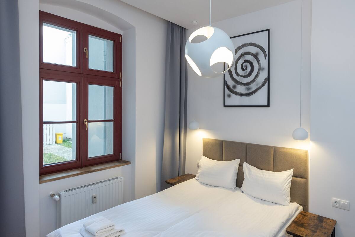 Un lit bien fait, avec du linge de lit blanc, une tête de lit en bois et une table de chevet sur laquelle se trouve une lampe. On aperçoit une fenêtre avec un cadre rouge et des rideaux, et un tableau abstrait encadré est accroché au mur.
