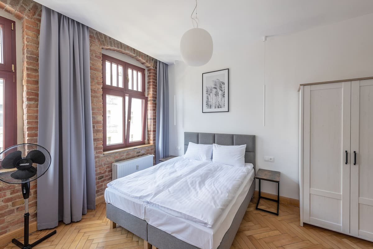 Une chambre avec un mur en briques, une grande fenêtre avec des rideaux, un lit avec une literie blanche, un ventilateur sur pied et une armoire.