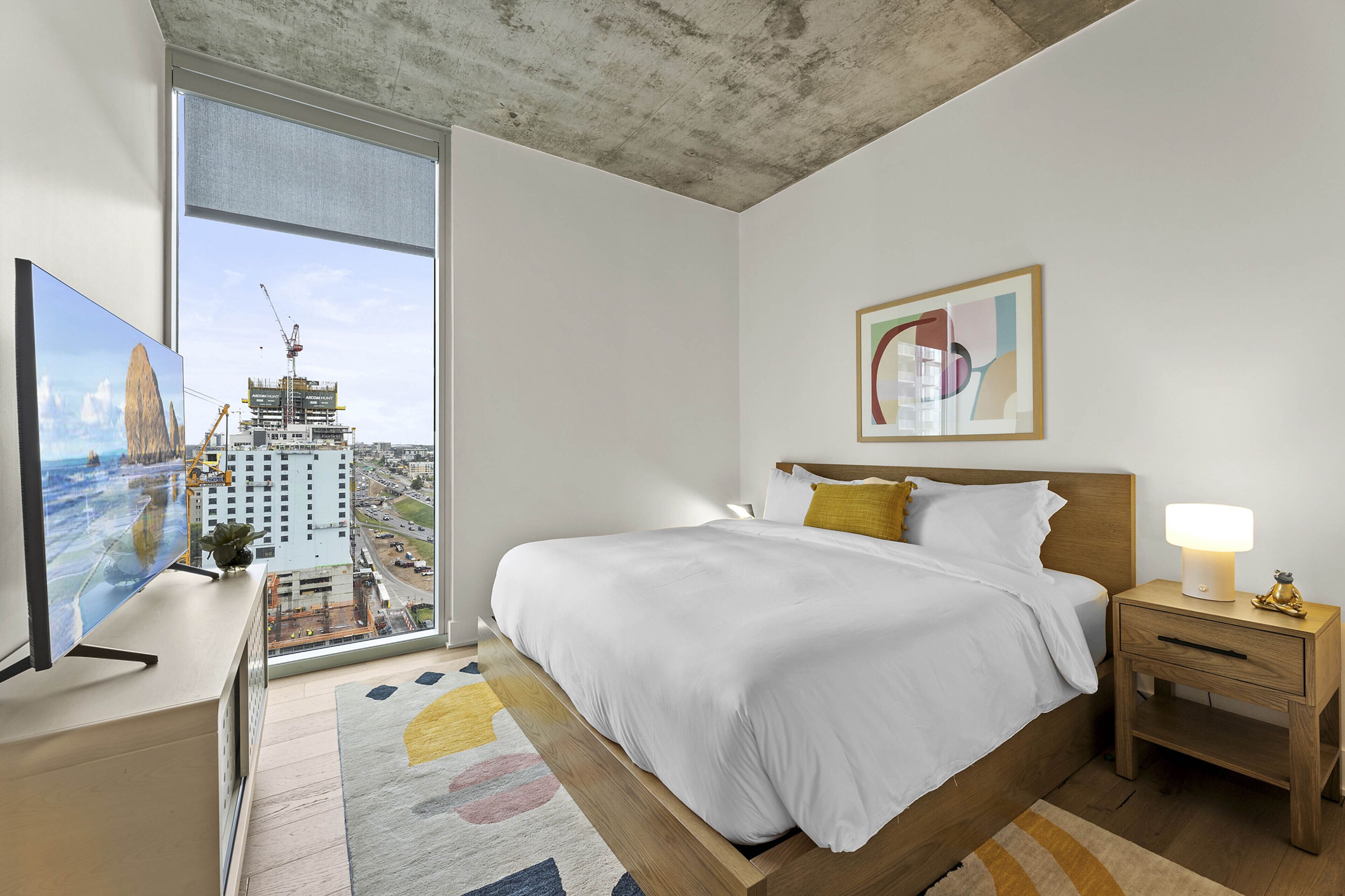 A modern hotel room with a large window offering a city view, a bed with white bedding, a bedside table with a lamp, and a painting on the wall.