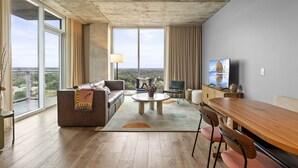 A spacious living room with a large window, a sofa, a coffee table, and a dining area. - Luxe Rainey St High Rise | Heated Pool | Gym | ATX (Austin)