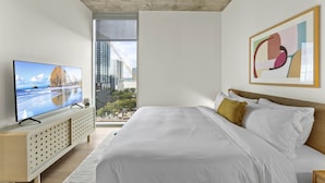 A modern hotel room with a large bed, a television on a low cabinet, and a city view through the window. - Luxe Rainey St High Rise | Heated Pool | Gym | ATX (Austin)
