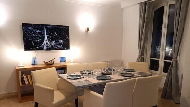 A dining area with a table set for four, a television mounted on the wall, and a bookshelf with books. - Aramis Colibri 72m2, lit King-size, Baignoire duo (Paris)