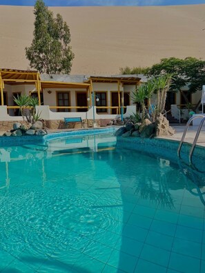 Una piscina con agua azul cristalina, rodeada por un edificio con barandillas y ventanas amarillas, y un paisaje desértico al fondo.