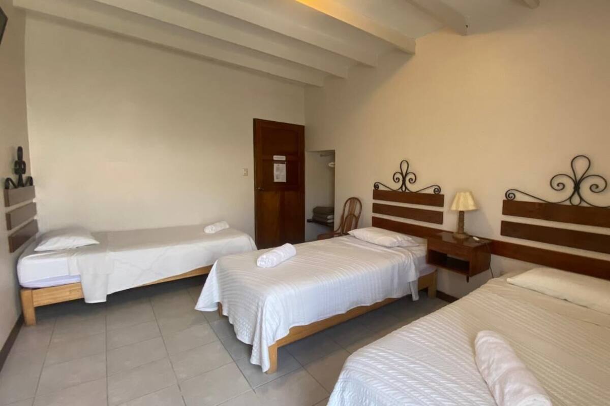A hotel room with three single beds, each with a wooden headboard and white bedding. There is a wooden nightstand with a lamp between the beds.