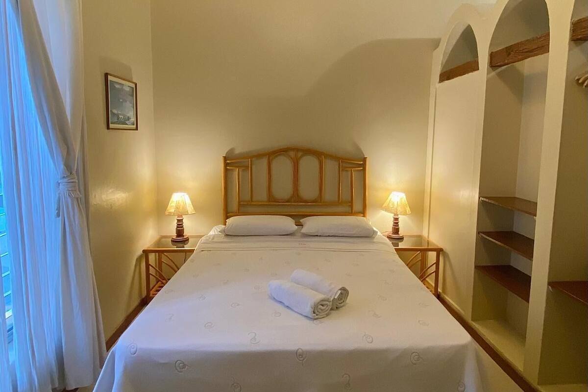 A neatly made bed with a wooden headboard, two bedside lamps, and a rolled towel on the bed.