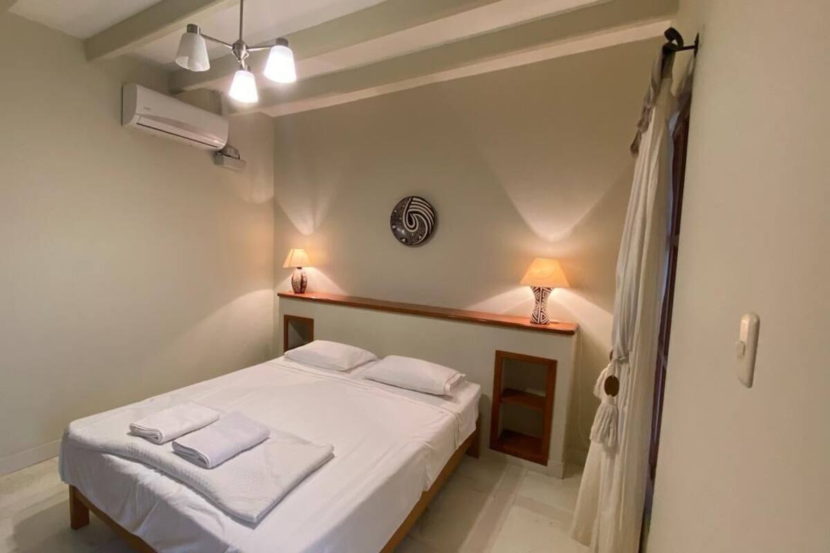 A neatly made bed with white linens and towels, a bedside table with a lamp, and a wall-mounted air conditioner.