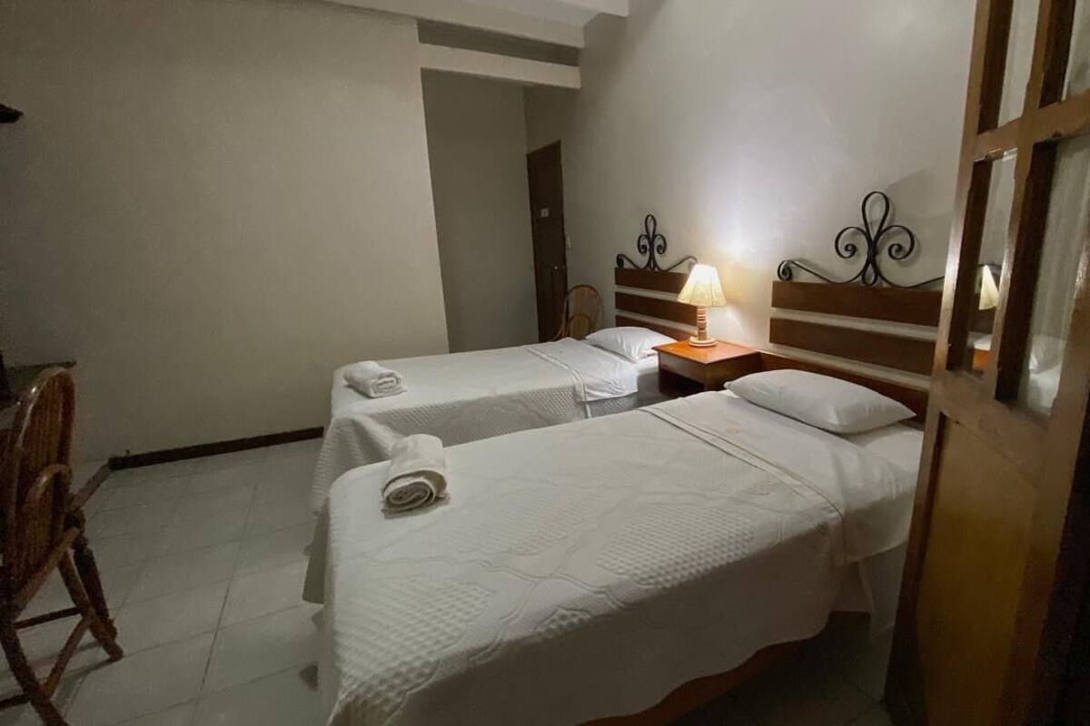 A room with two single beds, a wooden nightstand with a lamp, a chair, and a door with a glass panel.