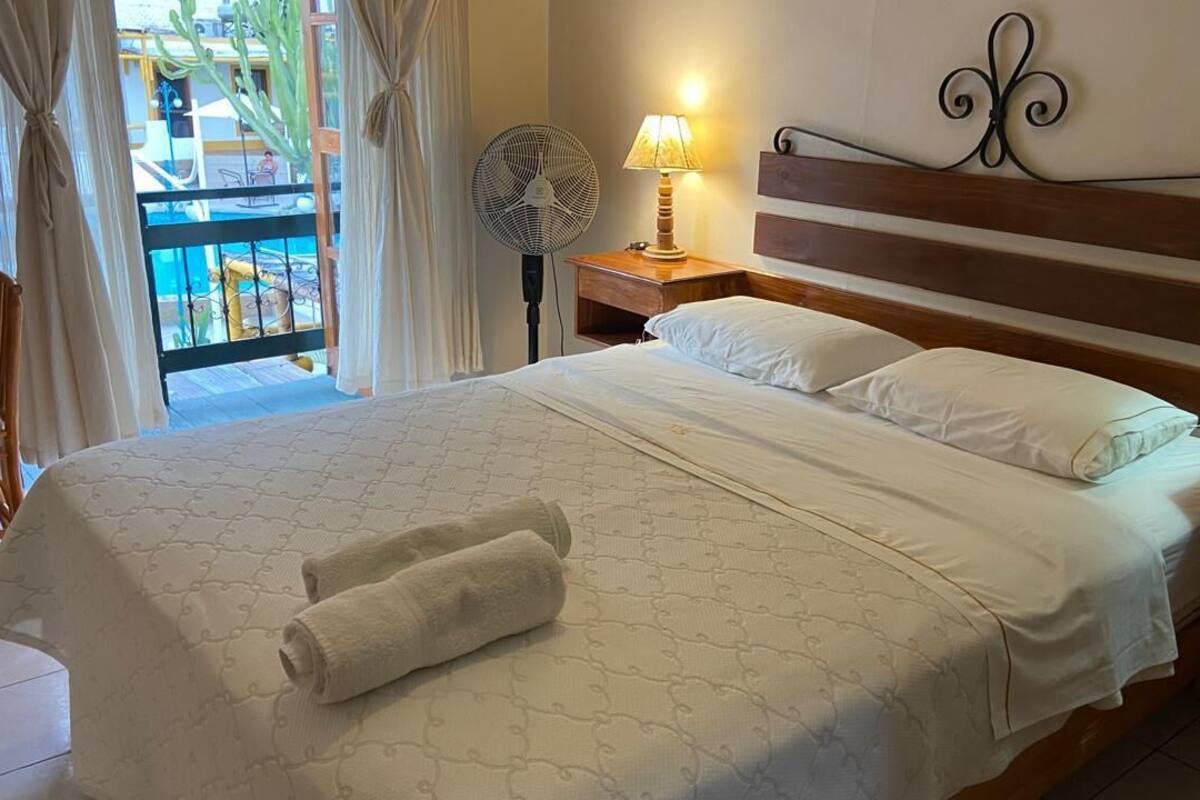 A hotel room with a large bed, a bedside table with a lamp, a fan, and a view of a pool area.