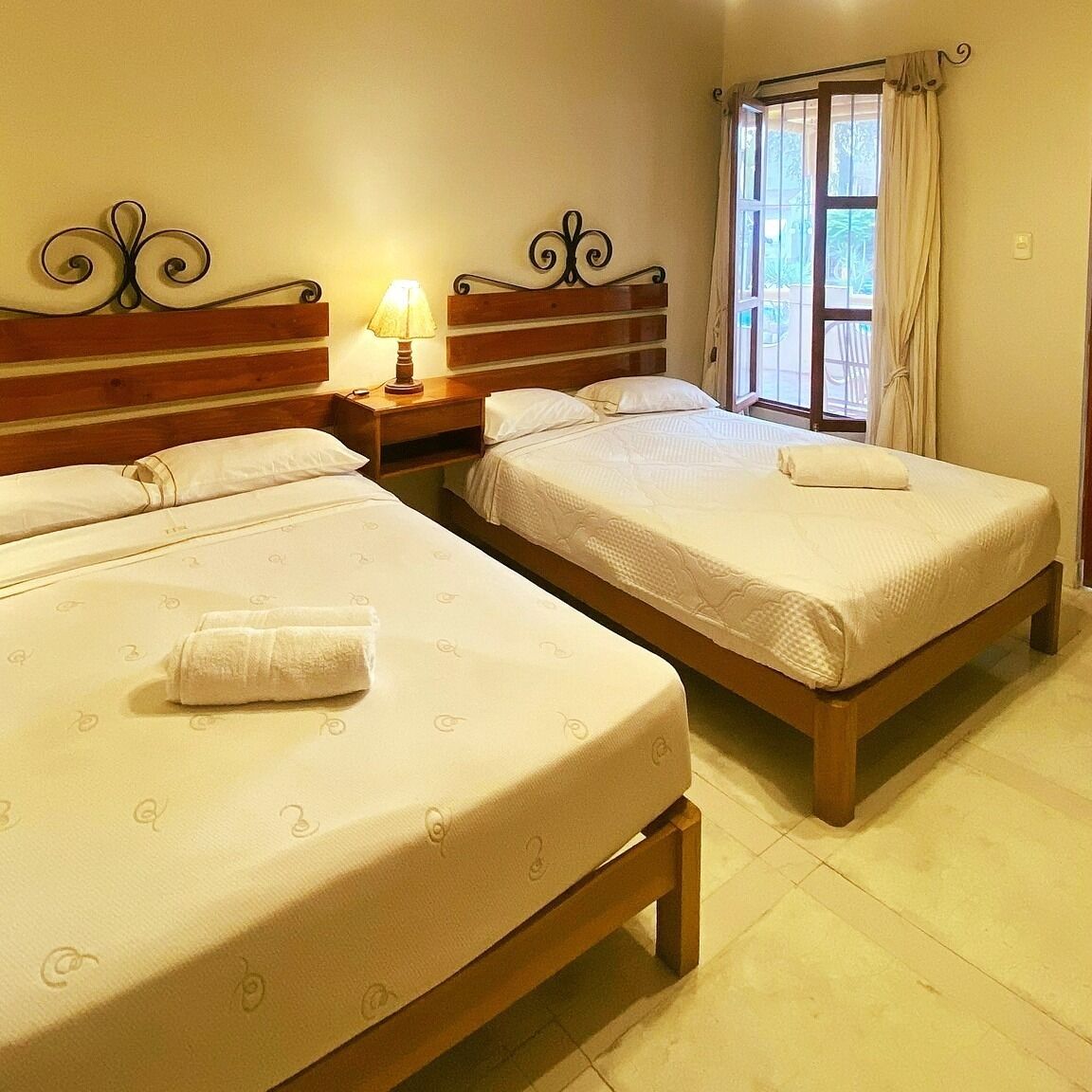 Habitación de hotel con dos camas individuales, una mesita de noche con lámpara y una ventana con cortinas.