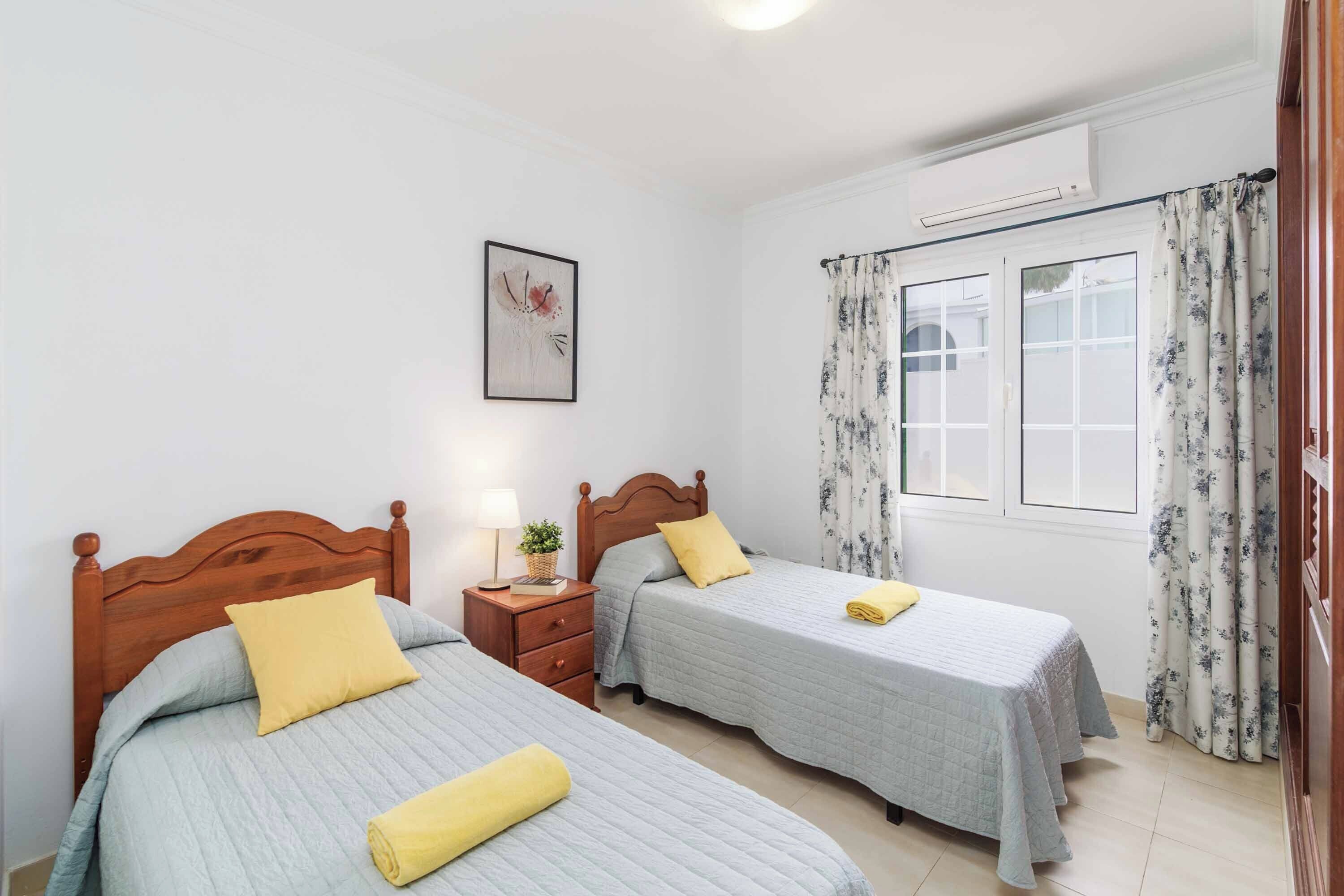 A bedroom with two beds, a nightstand, a window with curtains, and a wall-mounted air conditioner.